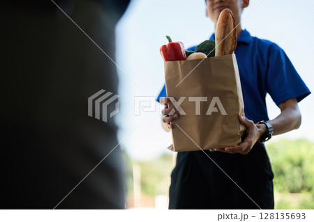 Delivery Service Concept - Close-Up of a Delivery Person Handing Over a Paper Bag of Groceries to a Customer at the Doorstep, Representing Modern Convenience and Efficient Service Delivery Service Concept - Close-Up of a Delivery Person Handing Over a Paper Bag of Groceries to a Customer at the Doorstep, Representing Modern Convenience and Efficient Service 128135693