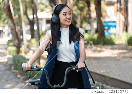 Young Woman Enjoying an Eco-Friendly Lifestyle, Riding a Bicycle in a Modern Urban Park, Embracing Sustainable Living with a Smile, Wearing Casual Clothing and Headphones, Surrounded by Nature 128135773