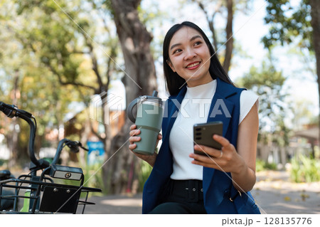 Young Professional Embracing Eco-Friendly Lifestyle with Reusable Cup and Smartphone in Urban Park Setting, Promoting Sustainable Business Practices and Modern Eco-Conscious Living 128135776