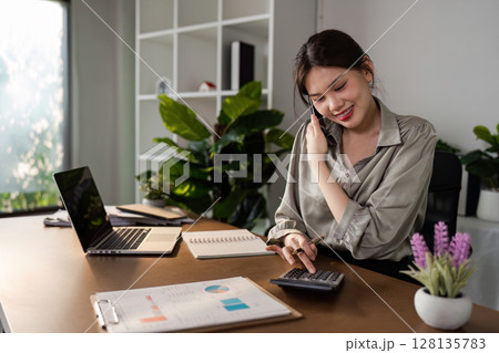 Young Business Professional Woman Working from Home Office, Smiling and Using Calculator, Surrounded by Modern Office Supplies, Laptop, and Indoor Plants, in a Bright and Minimalist Workspace 128135783