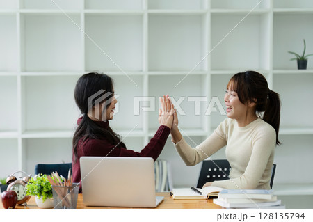 Two Business Women High-Fiving in Modern Office Setting with Laptop and Books, Celebrating Success and Teamwork, Smiling and Happy, Bright and Minimalist Workspace with Shelves in Background 128135794