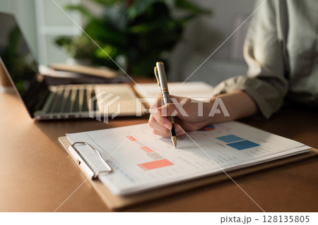 Business Professional Analyzing Financial Data on a Clipboard with Laptop in Background in Modern Office Setting 128135805