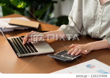 Professional Businesswoman Working on Laptop and Calculator in Modern Office Setting with Documents and Charts 128135806