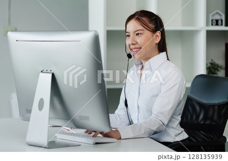 Professional Asian Woman Working at Computer with Headset in Modern Office Environment, Smiling and Engaged in Customer Service or Online Communication 128135939