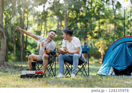 Couple gay lgbt Enjoying a Camping Trip in the Forest, Relaxing in Camping Chairs with a Tent in the Background, Sharing a Laugh and Drinking Coffee, Embracing the Outdoors and Nature 128135978