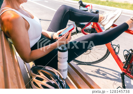 Cyclist Woman Resting on Bench with Smartphone, Red Bicycle and Helmet on a Sunny Summer Day Cyclist Woman Resting on Bench with Smartphone, Red Bicycle and Helmet on a Sunny Summer Day 128145738