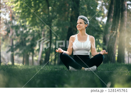 Smiling Mature Woman Meditating Outdoors in Sukhasana Yoga Pose Surrounded by Nature 128145761