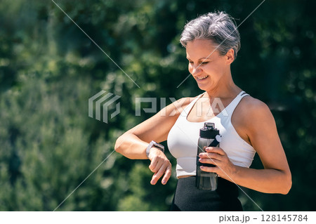 Mature Fit Woman Checking Smartwatch During Outdoor Workout Holding Water Bottle in Hand Mature Fit Woman Checking Smartwatch During Outdoor Workout Holding Water Bottle in Hand 128145784