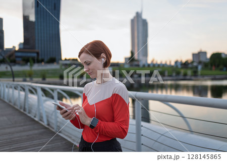 Runner checking her performance on a smartphone during her morning run across a city bridge. 128145865