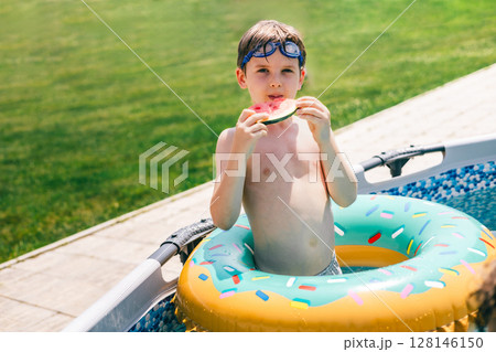 Cute boy in inflatable ring eating watermelon by backyard pool on a bright and sunny summer day 128146150