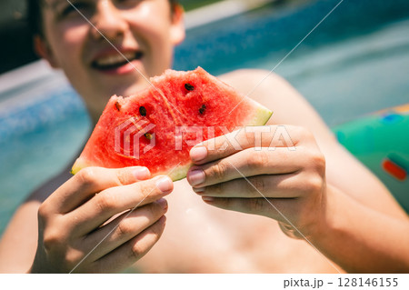 Teen boy holding juicy watermelon slice at poolside on sunny summer day, close-up of hands 128146155