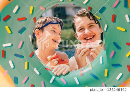 Two cheerful boys sharing watermelon through colorful pool float on sunny summer afternoon 128146162