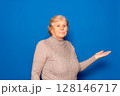 Caucasian woman in her 70s showing open palm offering new medical beauty procedure, anti-aging product, posing isolated over blue studio background, copy space for advertising text concept. 128146717