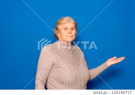 Caucasian woman in her 70s showing open palm offering new medical beauty procedure, anti-aging product, posing isolated over blue studio background, copy space for advertising text concept. 128146717