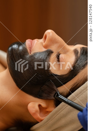Woman relaxing with black charcoal face mask applied by brush during spa facial treatment Woman relaxing with black charcoal face mask applied by brush during spa facial treatment 128147540