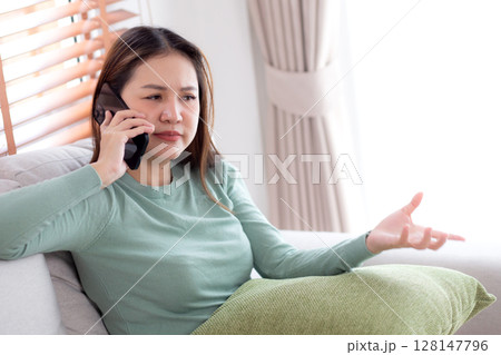 Asian young woman stress and worried sitting on sofa while speaking on smart phone in living room. 128147796