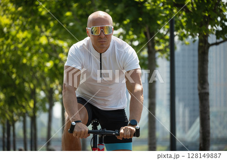 Caucasian cyclist in motion in white t-shirt rides bicycle through urban public park. Sporty man wears reflective sunglasses. Outdoor activity. Caucasian cyclist in motion in white t-shirt rides bicycle through urban public park. Sporty man wears reflective sunglasses. Outdoor activity. 128149887