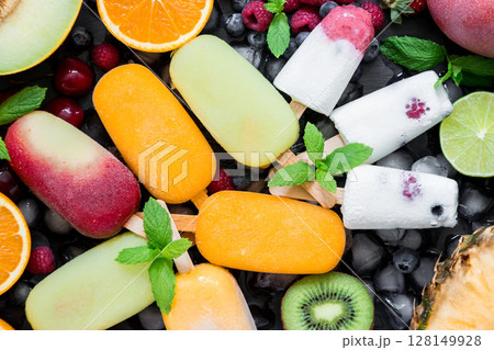 A vibrant array of berry flavored ice cream bars laid out flat, surrounded by slices of fresh fruits including melons, oranges, kiwis, and mint leaves for decoration. 128149928