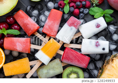 Gathered on a slate surface, a vibrant selection of berry ice cream popsicles lies flat, surrounded by fresh fruits and mint leaves. 128149933