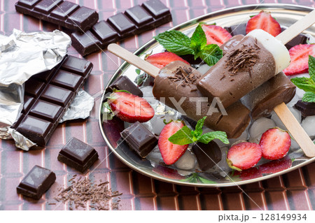 Chocolate vanilla ice cream on a silver platter with ice and chocolate pieces, mint, strawberry near pieces of chocolate on a chocolate background. Chocolate vanilla ice cream popsicles and stawberry. 128149934