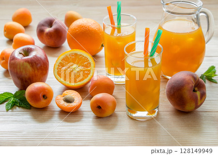 Fruit juice in glasses with ice near the jar of juice and scattered apricots, peaches, apple, orange on a light wooden background. Apricot peach apple orange juice with ice. Horizontal. 128149949