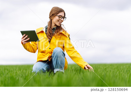 Young farmer woman in a green field, intently using a tablet. She wears a yellow jacket and glasses 128150694