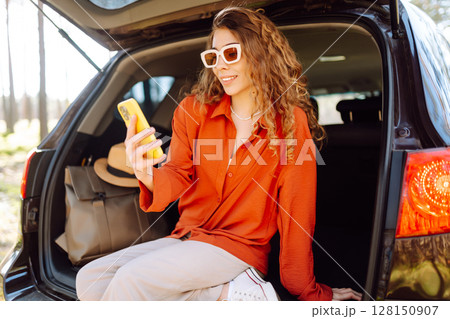 Beautiful woman sits in the trunk of a car outdoor and uses a smartphone. Lifestyle Beautiful woman sits in the trunk of a car outdoor and uses a smartphone. Lifestyle 128150907