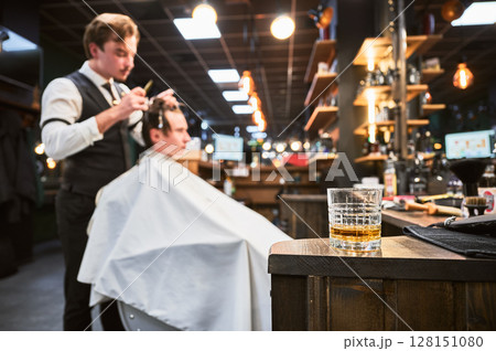Close-up photo of a glass of whiskey. High-quality barbershop offering drinks to customers. Glass with alcohol on the background of hairstylist making haircut. Close-up photo of a glass of whiskey. High-quality barbershop offering drinks to customers. Glass with alcohol on the background of hairstylist making haircut. 128151080