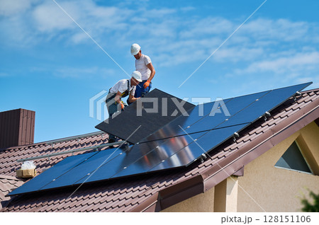 Workers building solar panel system on rooftop of house. Two men installers in helmets installing photovoltaic solar module outdoors. Alternative, green and renewable energy generation concept. 128151106