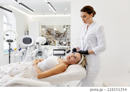 Cosmetologist using skincare equipment for photorejuvenation facial treatment. Mature woman lying on chair in beauty clinic before non-invasive anti-aging procedure with aesthetic laser technology Cosmetologist using skincare equipment for photorejuvenation facial treatment. Mature woman lying on chair in beauty clinic before non-invasive anti-aging procedure with aesthetic laser technology 128151354