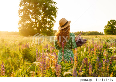 Stylish woman in a straw hat with a bouquet of flowers on a field with lupins. Lifestyle concept. 128151715