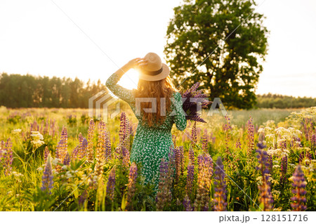 Stylish woman in a straw hat with a bouquet of flowers on a field with lupins. Lifestyle concept. 128151716