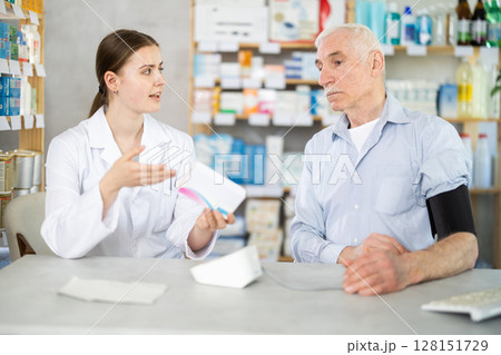Young female pharmacist offering pills to mature man having blood pressure measured Young female pharmacist offering pills to mature man having blood pressure measured 128151729