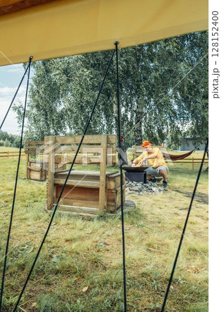 man in yellow near outdoor fire pit surrounded by stones with rustic benches seen through tent ropes, camping equipment, travel agency, outdoor adventure company, vertical man in yellow near outdoor fire pit surrounded by stones with rustic benches seen through tent ropes, camping equipment, travel agency, outdoor adventure company, vertical 128152400