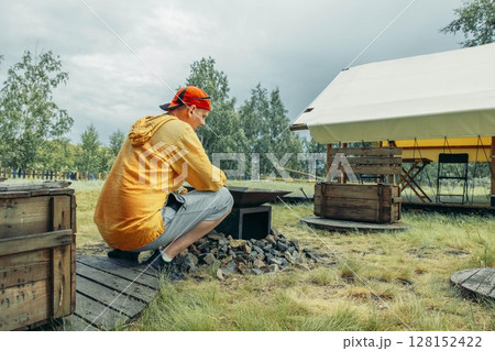 man in yellow squatting near outdoor fire pit surrounded by rocks with glamping tent in grassy field with trees, camping equipment, travel agency, outdoor adventure company, clouds 128152422