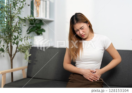Health Awareness: Young woman experiencing abdominal pain while sitting on a couch. Health Awareness: Young woman experiencing abdominal pain while sitting on a couch. 128153104