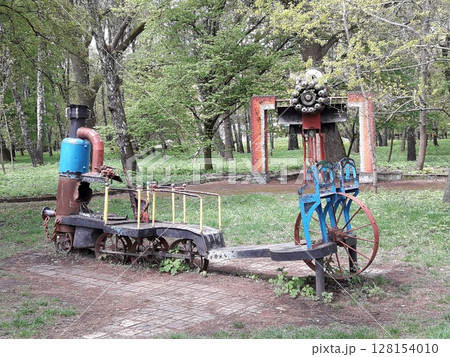 An Artistic Sculpture Resembling a Train with Wheels and a Chimney in a Park Among Trees An Artistic Sculpture Resembling a Train with Wheels and a Chimney in a Park Among Trees 128154010