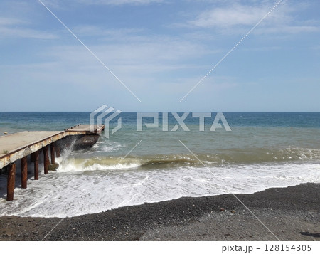 A tranquil beach scene with a weathered wooden pier extending into the sea, waves crashing against the dark pebbled shore under a clear blue sky 128154305