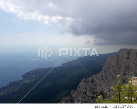 A dramatic view from a cliffside overlooking a lush green valley, a coastal town along the shoreline, and the vast blue sea under a partly cloudy and overcast sky 128154310