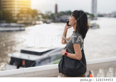 Content solo southeast asian woman teenager blogger on travel tourism in Bangkok Thailand. An explorer wait for local boat on Chao Phraya river in Asia using smartphone 128154453