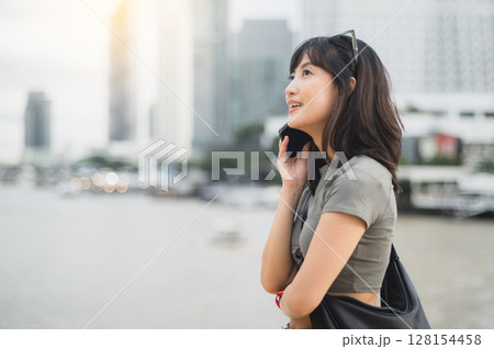 Happy solo Southeast Asian woman teenager blogger on tourism trip. Wait to express joy to friend with smartphone for boat on Chao Phraya river in Bangkok Thailand 128154458
