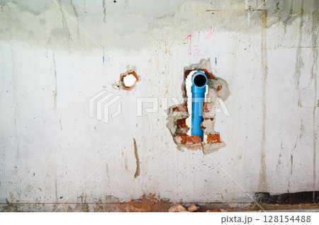 Damaged concrete wall with an exposed blue pipe in hole during major house renovation. Plumbing system work part of construction project with messy background view Damaged concrete wall with an exposed blue pipe in hole during major house renovation. Plumbing system work part of construction project with messy background view 128154488