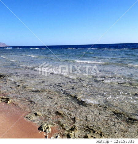 A picturesque view of a clear water beach with gentle waves lapping against the shore, surrounded by rocky formations under a bright blue sky A picturesque view of a clear water beach with gentle waves lapping against the shore, surrounded by rocky formations under a bright blue sky 128155333