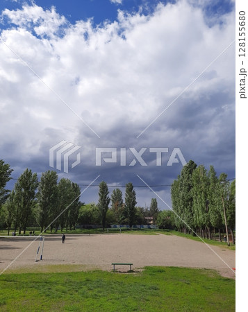 A wide open sports field with a sandy center, framed by tall green trees under a dramatic sky with towering white clouds and hints of blue, creating a striking natural scene 128155680