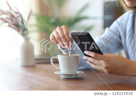 Woman hand using phone stirring coffee at home 128157019