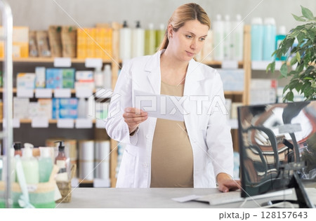 Pregnant woman employee in white coat works in pharmacy sales area using computer. 128157643