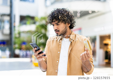 A man looking concerned while holding a credit card and smartphone in a shopping mall setting. 128157655