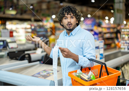 A shocked man looks at his grocery bill, holding a shopping basket in a supermarket. He is surprised by the total. A shocked man looks at his grocery bill, holding a shopping basket in a supermarket. He is surprised by the total. 128157656