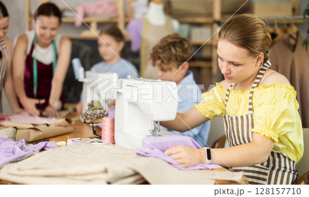Teenage girl works on sewing machine while teacher and children learn how to cut fabric 128157710