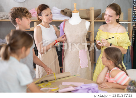 Female dressmaker teacher shows children how to try on dresses on mannequin in classroom 128157712